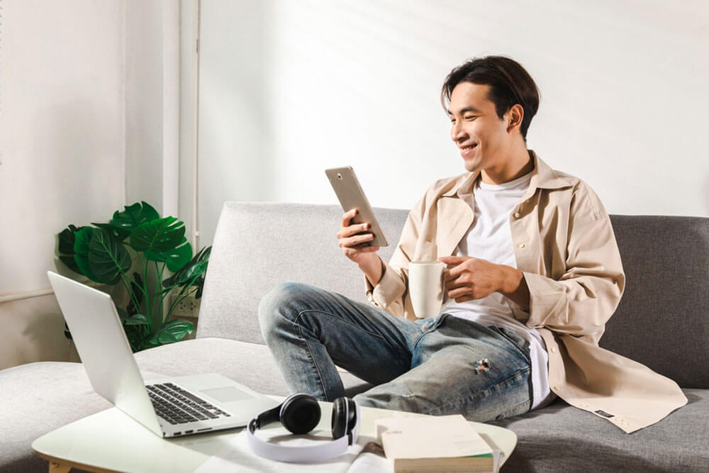 Happy smiling Asian student sits and relaxes on a couch using modern table